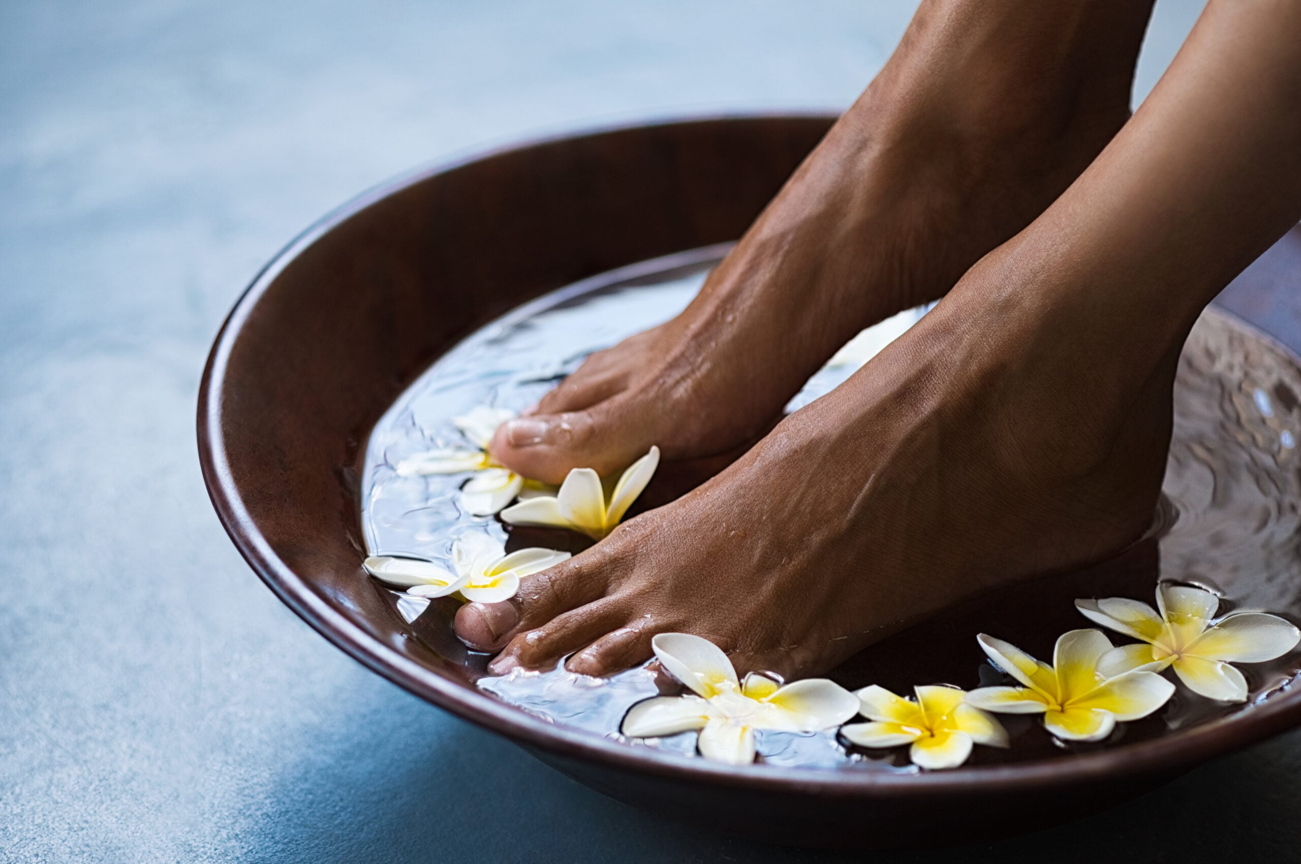 Woman soaking feet in bowl of water with floating frangipani flowers at spa. Closeup of a female feet at wellness center on pedicure procedure. Woman feet in spa wooden bowl with exotic white flowers.